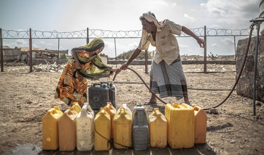 A father and daughter stand in front of a row of yellow water containers, filling them up from a nearby water access station.