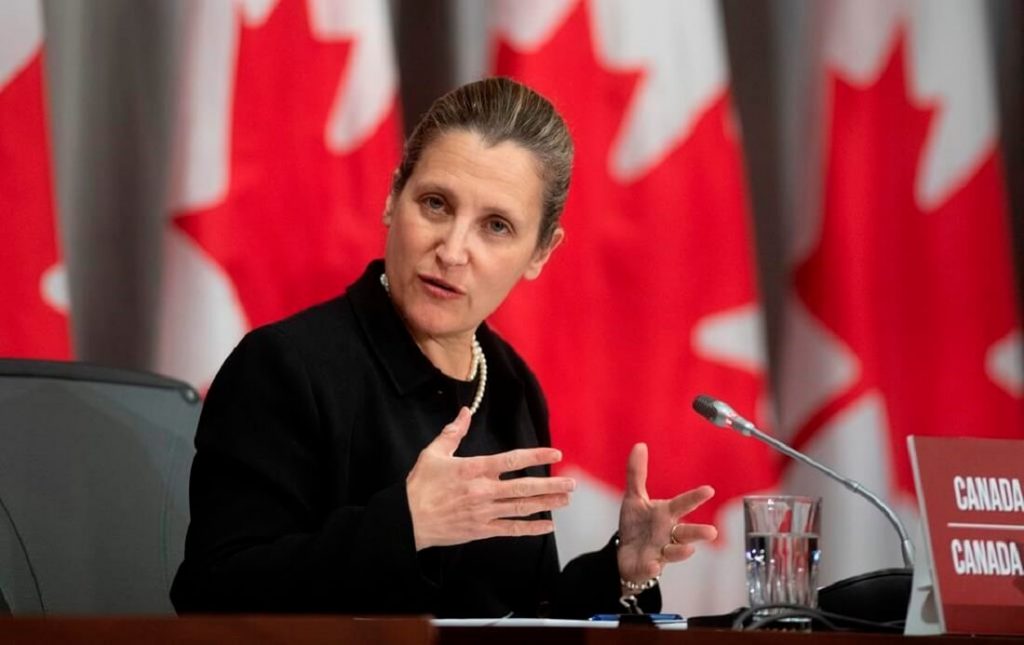 Canada's Finance Minister Chrystia Freeland is gesturing with her hands and speaking into a microphone at a public speaking event. There are several Canadian flags behind her.