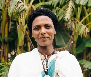 A woman in white clothes standing in front of trees while looking at the camera with an optimistic expression