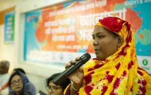 A woman wearing a red and yellow headscarf speaking to a group of women using a microphone