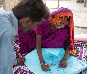 A woman wearing bright purple, sitting on the floor with a man and drawing instructions on a large piece of light blue paper