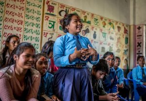 A young woman in a blue school uniform, presenting something to her classmates